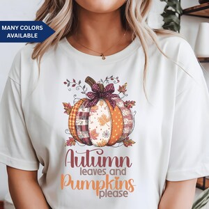May include: Cream-colored t-shirt featuring a colorful pumpkin design with plaid, polka dot, and leaf patterns. The pumpkin has a brown stem and a burgundy bow. The text "Autumn leaves and pumpkins please" is below the pumpkin.