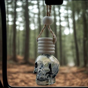 Puede incluir: Un ambientador para coche transparente con forma de calavera, con un anillo de madera y un colgador de cuerda. El ambientador está suspendido contra un fondo borroso de bosque. El diseño de calavera añade un toque único.