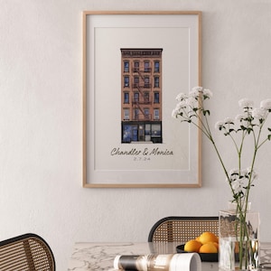 May include: A framed wall art print featuring a watercolor illustration of a brownstone building with a fire escape. Below the building, the text reads Chandler & Monica and the date 2.7.24. The print is displayed in a light wood frame.