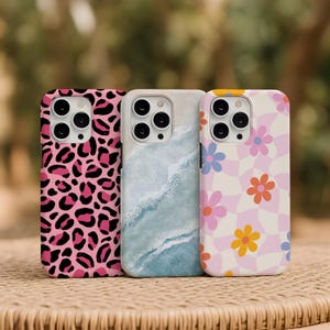 May include: Three phone cases with unique designs. The first case has a pink leopard print, the second has a blue and white watercolor design, and the third features a pink and white checkered pattern with colorful flowers.