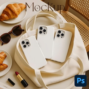 May include: A cream-colored tote bag holds three white phone cases. A croissant sits on a white plate, and a pair of brown sunglasses rests nearby. A tube of lipstick and a black lipstick cap are also visible.