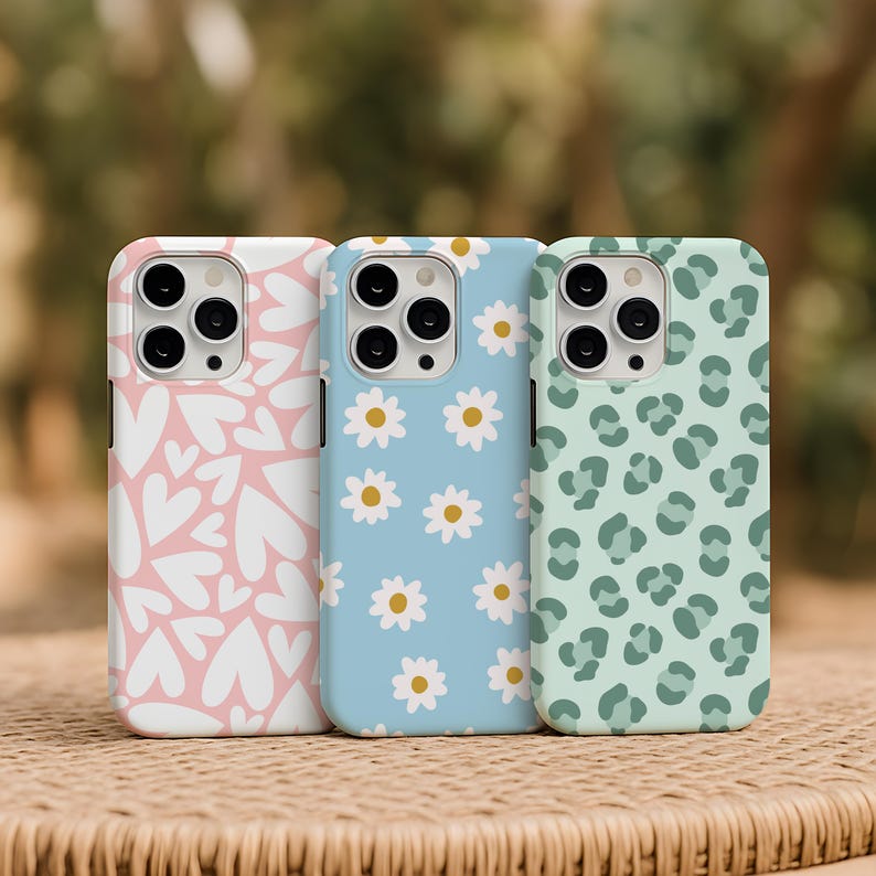 May include: Three phone cases with different designs. One is light pink with white hearts, another is light blue with white daisies, and the third is light green with a leopard print pattern. The cases are on a woven surface.