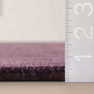 以下が含まれることがあります： 紫色のカーペットの一部のクローズアップ。カーペットは厚く、質感のあるパイルです。定規がカーペットの端に沿って垂直に配置され、センチメートルを示すマークが付いています。カーペットの色は、深く落ち着いた紫色です。
