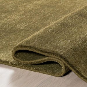 Op de afbeelding: Een effen olijfgroen vloerkleed met een dichte, laagpolige textuur. Het kleed is op één hoek omgevouwen, waardoor de dikte van het materiaal en de consistente, diepgroene kleur over het gehele oppervlak van de vloerbedekking zichtbaar zijn.