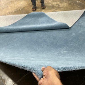May include: A large, rectangular area rug featuring a solid, muted blue top surface with a dense, soft-textured pile. The rug is partially folded back to reveal a light grey, smooth, and flat backing material on the underside of the textile.
