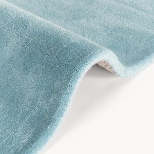 May include: Close-up of a section of a light blue rug. The rug has a soft, plush texture and a slight sheen. The edge of the rug is rolled over, revealing a beige backing. The color is a muted, calming blue.