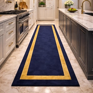 May include: A rectangular navy blue rug with a golden yellow border. The rug is placed in a kitchen with white cabinets, a stainless steel oven, and a marble countertop. The rug's dimensions are not visible.