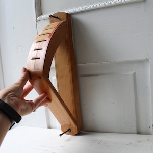 Op de afbeelding: Een houten wandgarenklos, een hulpmiddel om garen tot een bol te wikkelen. De klos is gemaakt van lichtgekleurd hout met een gebogen ontwerp en verschillende sleuven. Het heeft een rood geschilderd accent. Een persoon houdt de klos vast.