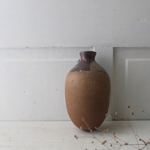May include: A brown ceramic vase with a dark brown rim, sitting on a white surface. The vase is slightly wider at the bottom and tapers towards the top.