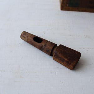May include: A vintage wooden bottle stopper with a dark brown finish. The stopper has a cylindrical body with a hole and a rectangular end. The wood shows signs of wear, suggesting age and use.