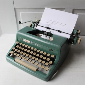 May include: Vintage green Royal typewriter with off-white keys and a paper sheet inserted. The typewriter has a classic design with a metal body and a beige space bar. The paper shows typed text.