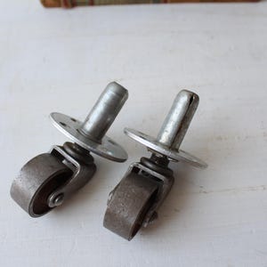 May include: Two antique metal caster wheels with a silver finish. Each caster has a small wheel, a spring, and a mounting plate with screw holes. The casters are designed for furniture or other items needing mobility.