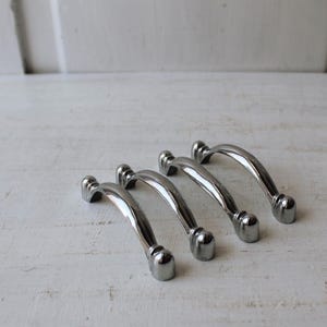 May include: Four polished silver cabinet pulls with a curved design. Each pull has rounded ends and a smooth, reflective surface. The pulls are arranged on a white, textured surface, suggesting a hardware or home decor theme.
