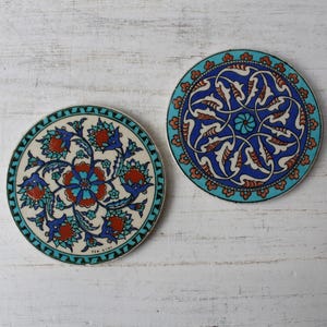 May include: Two circular ceramic coasters featuring intricate patterns. The left coaster has a cream background with red, blue, and green floral motifs. The right coaster displays a blue and turquoise geometric design with a central flower.