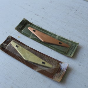 May include: Two metal corner brackets in their original packaging. The brackets are in the shape of a triangle with rounded corners. One bracket is gold and the other is copper.