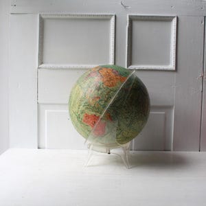 May include: A spherical globe featuring a detailed map with green landmasses and orange highlighted regions, mounted on a clear, transparent acrylic stand that curves around the side of the sphere to provide support and stability.