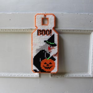 May include: A handmade Halloween door hanger featuring a witch with a black hat and a jack-o'-lantern. The word "BOO!" is at the top. The door hanger is made of orange, black, white, and green yarn.
