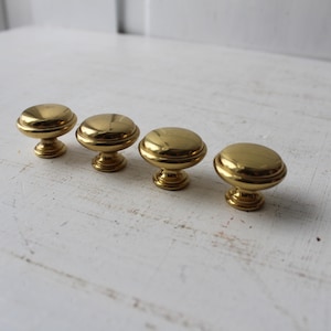May include: Four brass cabinet knobs with a rounded top and a decorative base. The knobs are golden in color and are arranged in a row on a white surface. Each knob measures approximately 1.25 inches in diameter.