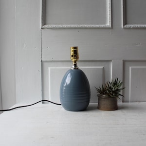 May include: A smooth, egg-shaped table lamp base in a dusty blue finish with horizontal ribbed detailing near the bottom. The lamp features a metallic gold-toned socket at the top and includes a black electrical cord extending to the left side.