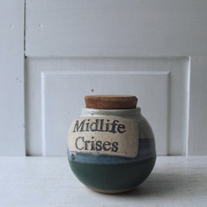 May include: A small, handcrafted ceramic jar with a cork stopper. The jar is painted in shades of green, blue, and beige, with the words "Midlife Crises" printed on a label. The jar is sitting on a white surface.