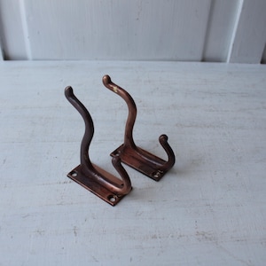 May include: Two antique bronze-colored metal wall hooks. Each hook has a rectangular base with a curved, decorative hook. The hooks are a dark brown color with a slight sheen, suggesting an aged finish. They are mounted on a white surface.