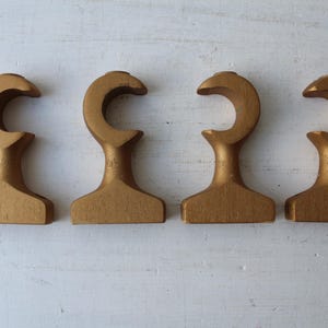 May include: Four gold-colored wooden hooks arranged in a row. Each hook has a curved top and a rectangular base. The hooks are displayed on a white, textured surface.