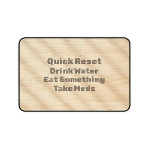 May include: A rectangular wooden desk mat with rounded corners. The mat features the text "Quick Reset Drink Water Eat Something Take Meds" in a gray, textured font. The wood grain pattern is visible.
