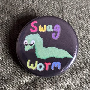 May include: A circular, black badge featuring the words "Swag Worm" in colourful bubble letters. A green worm wearing sunglasses is in the centre of the badge.