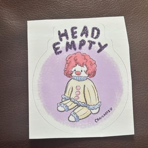 May include: A sticker depicting a cartoon clown with a red afro, in a yellow outfit with blue trim and pink buttons. The text "HEAD EMPTY" is above the clown, with "CRISISARTZ" written below.