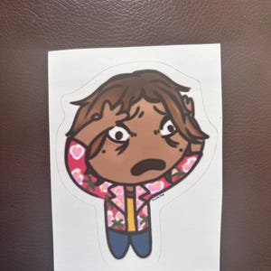 Peut inclure: Un autocollant représentant un personnage de dessin animé à la peau brune, aux cheveux bruns et aux yeux écarquillés, exprimant la surprise. Le personnage porte une veste rose à imprimé floral, une chemise jaune et un pantalon bleu. L'autocollant est sur fond blanc.