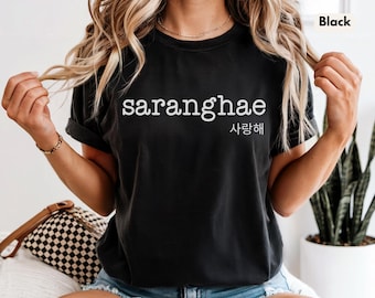 Puede incluir: Camiseta negra con la palabra "saranghae" en blanco, con caracteres coreanos debajo. La camiseta es de manga corta. La persona lleva una pulsera dorada y vaqueros azules. La camiseta forma parte de una colección de ropa.