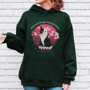Puede incluir: Sudadera con capucha verde oscuro con un gráfico de un gato blanco y negro dentro de un círculo rosa, rodeado de flores de cerezo. El texto en la sudadera dice "Main Lead Energy: Goyangi Edition" y "Petals, Purrs, and Plot Twists."