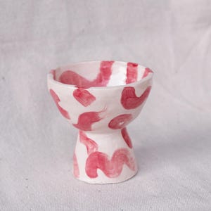 May include: A small, white ceramic bowl with a pedestal base. The bowl is decorated with abstract red brushstrokes and shapes. The interior and exterior of the bowl are painted with the same design. The bowl is approximately 3 inches tall.