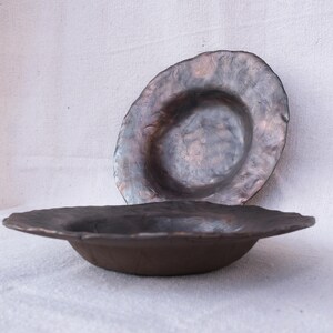 May include: Two handmade, rustic ceramic bowls. The bowls are a dark brown color with a textured surface and irregular edges. One bowl is in the foreground, and the other is behind it. The bowls are on a white surface.