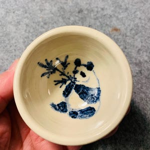 Puede incluir: Pequeño cuenco de cerámica blanquecina con un diseño de oso panda pintado a mano en azul. El panda sostiene bambú. El cuenco es redondo y tiene una estética sencilla y minimalista. Ideal para servir pequeñas porciones o como pieza decorativa.