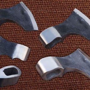 May include: Close-up of several axe heads with a textured, dark gray finish on the blade and a shiny, silver-colored poll. The axes are arranged on a brown, textured surface.