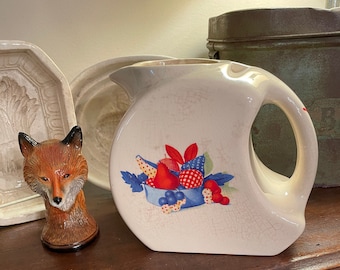 May include: Cream-colored vintage pitcher with a fruit design. The pitcher features a large handle and a spout. The fruit design includes red, blue, and white elements. A ceramic fox figurine and other decorative items are in the background.
