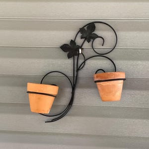 May include: A wall-mounted plant holder featuring a black metal frame with decorative scrollwork and two leaf-shaped accents. The frame supports two terracotta pots, each held securely within a circular metal ring, creating a symmetrical display.