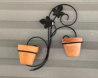 May include: A wall-mounted plant holder featuring a black metal frame with decorative scrollwork and two leaf-shaped accents. The frame supports two terracotta pots, each held securely within a circular metal ring, creating a symmetrical display.