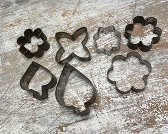 May include: A set of eight vintage metal cookie cutters in various shapes, including flowers, stars, and a spade. The cutters are on a white wooden surface.