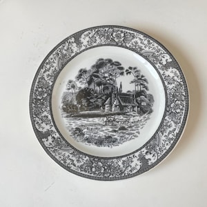 May include: A round white ceramic plate featuring a detailed black transferware print. The center depicts a rural landscape with a cottage, trees, and a horse-drawn carriage. The wide rim is decorated with a dense floral and geometric pattern.