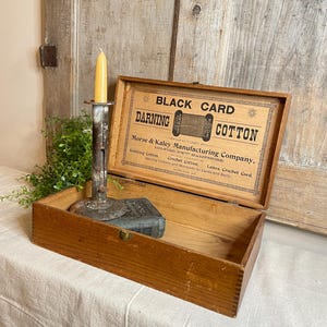 May include: An open wooden box with a hinged lid, displaying the text "BLACK CARD DARNING COTTON." Inside, a small book and a metal candle holder with a lit yellow candle are visible. The box rests on a white cloth.