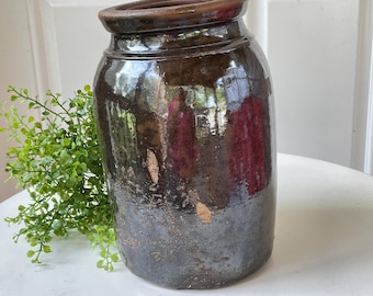 May include: A cylindrical stoneware crock with a dark brown, high-gloss glaze and a thick, rounded rim. The surface features visible texture and small areas of wear, showing a lighter brown base material beneath the dark, reflective finish.