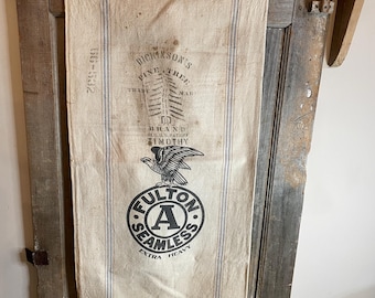 May include: Vintage, off-white fabric with blue vertical stripes and printed text. The text includes "DICKINSON'S PINE TREE BRAND" and "FULTON SEAMLESS EXTRA HEAVY" with an eagle emblem. The fabric is aged and mounted on a wooden door.