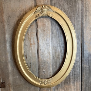 May include: Gold-colored oval picture frame with decorative elements. The frame features an eagle and banner at the top and a bow and arrow design at the bottom. The frame is made of a metallic material and has a smooth finish.