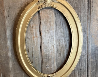 May include: Gold-colored oval picture frame with decorative elements. The frame features an eagle and banner at the top and a bow and arrow design at the bottom. The frame is made of a metallic material and has a smooth finish.