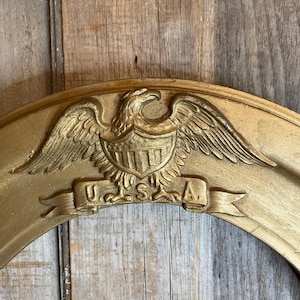 May include: A gold-colored, oval-shaped frame with an embossed eagle emblem and the letters "U.S.A." on a banner. The frame has a distressed finish, suggesting an antique or vintage style. The background is a wooden surface.