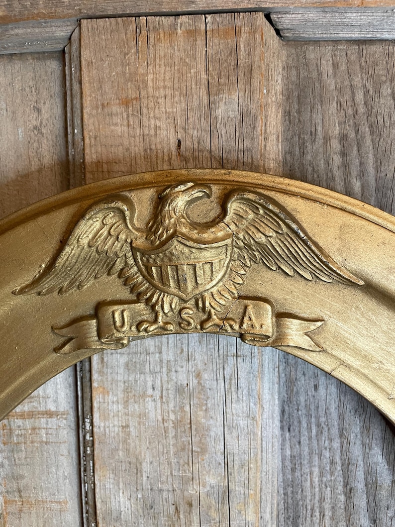 May include: A gold-colored, oval-shaped frame with an embossed eagle emblem and the letters "U.S.A." on a banner. The frame has a distressed finish, suggesting an antique or vintage style. The background is a wooden surface.