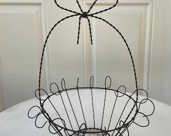 May include: A dark brown wire basket featuring a circular base with a grid pattern, a flared open top rim decorated with small wire loops, and a tall, arched handle that is twisted at the top to form a decorative bow shape.