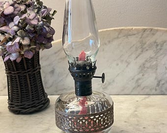 May include: A clear glass oil lamp featuring a rounded base with a decorative metallic band, a black burner assembly with a side adjustment knob, and a tall, tapered glass chimney. A pink wick is visible inside the burner and the glass base.